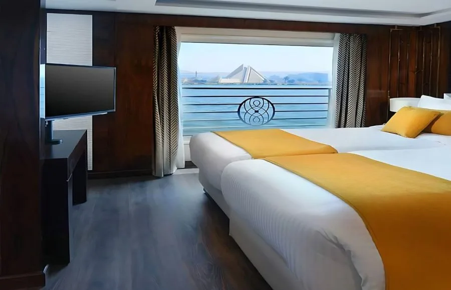 A comfortable cabin on the Sindbad Nile Cruise, featuring twin beds, a television, and a large window offering a stunning view of a felucca sailing the Nile.