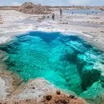 A mesmerizing Close photo of the deep turquoise water of a natural salt-ringed pool, part of the Siwa Salt Lake system in Egypt, with a tourist standing near the bank.