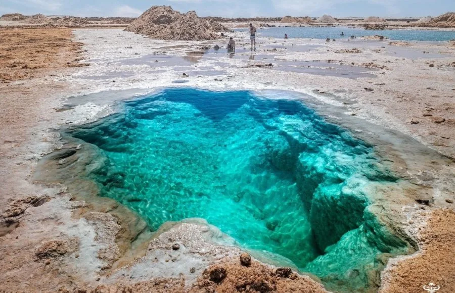 A mesmerizing Close photo of the deep turquoise water of a natural salt-ringed pool, part of the Siwa Salt Lake system in Egypt, with a tourist standing near the bank.