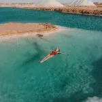 An aerial photo of a woman in a red swimsuit floating effortlessly on the vivid turquoise water of a Siwa Salt Lake in Egypt, with white salt deposits on the banks and large salt mounds in the background.
