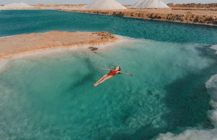 An aerial photo of a woman in a red swimsuit floating effortlessly on the vivid turquoise water of a Siwa Salt Lake in Egypt, with white salt deposits on the banks and large salt mounds in the background.