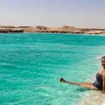 A Lady sits and relaxes at the edge of the vibrant turquoise waters of the Siwa Salt Lake in Egypt, enjoying a Relaxing View of the salt-encrusted shore.