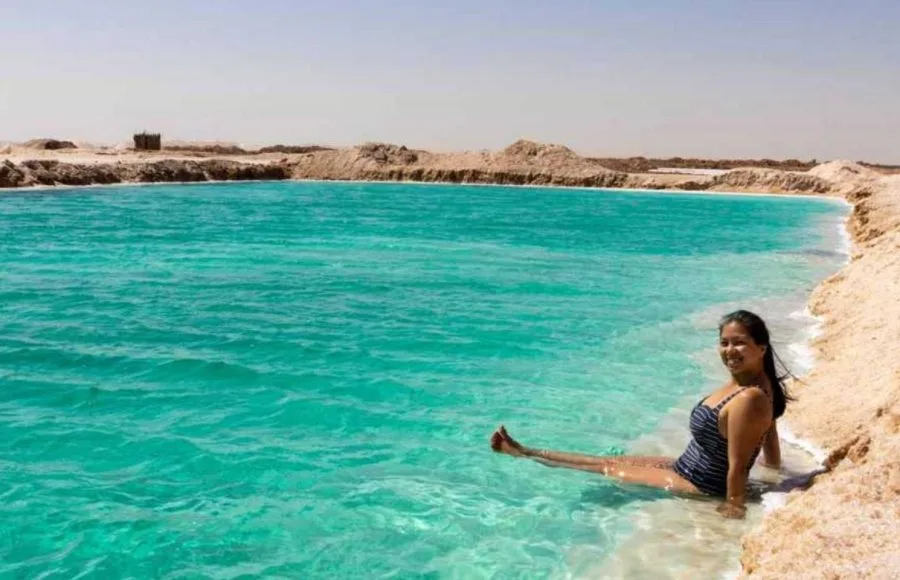A Lady sits and relaxes at the edge of the vibrant turquoise waters of the Siwa Salt Lake in Egypt, enjoying a Relaxing View of the salt-encrusted shore.