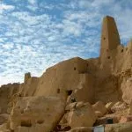 The ruins of Siwa The Temple Of The Oracle Amun Temple in Egypt, showing the ancient mud-brick walls and tapering tower against a bright blue sky with scattered clouds.