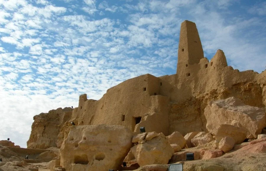 The ruins of Siwa The Temple Of The Oracle Amun Temple in Egypt, showing the ancient mud-brick walls and tapering tower against a bright blue sky with scattered clouds.