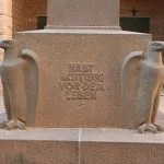 A close-up view of the stone base and eagle statues at The German War Memorial in El Alamein, featuring the German inscription, "HABT ACHTUNG VORDEM LEBEN" (Respect Life).