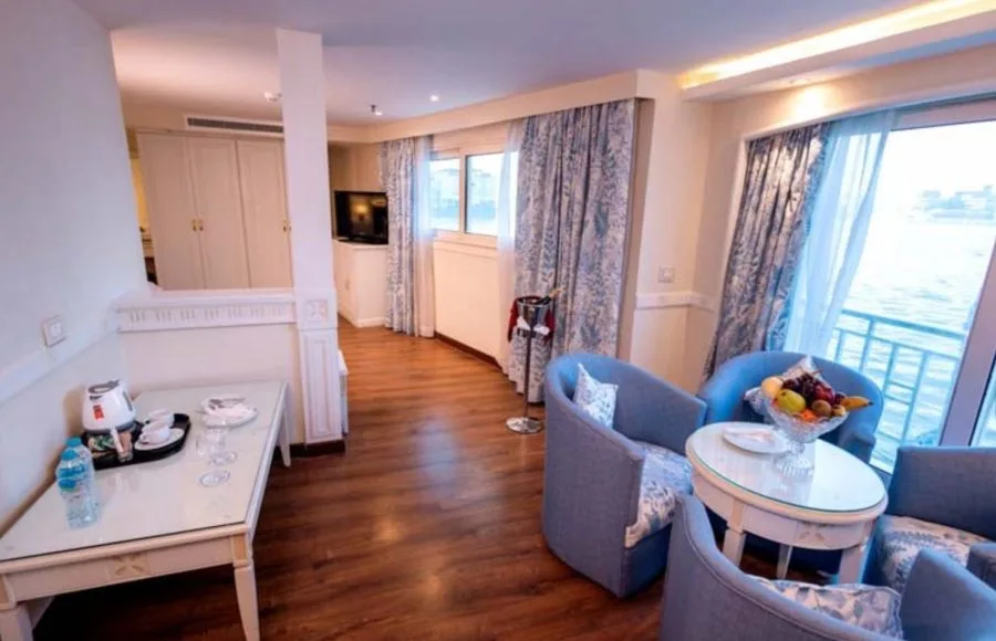 Luxurious cruise cabin lounge area with light purple armchairs, a side table, and a large window overlooking the water.