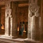 The interior hypostyle hall of the Abu Simbel Temple, showing large columns adorned with Hathor heads and tourists viewing the dimly lit, carved walls.