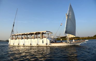 A traditional two-masted wooden sailboat, the Abundance Dahabiya, cruising the Nile River with open sails