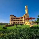 A beautiful, wide view of Al-Haramlik Montaza Palace in Alexandria, featuring the ornate yellow and red historical structure, a tall central tower, and lush green grass and palm trees in the expansive gardens.