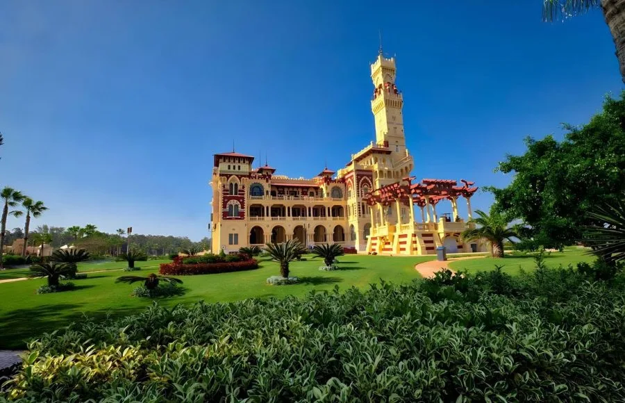 A beautiful, wide view of Al-Haramlik Montaza Palace in Alexandria, featuring the ornate yellow and red historical structure, a tall central tower, and lush green grass and palm trees in the expansive gardens.
