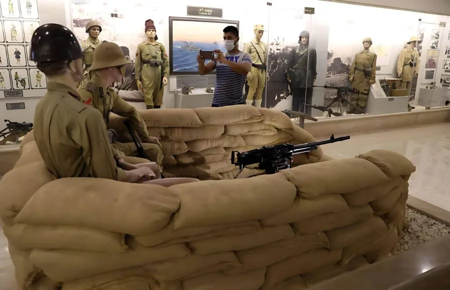 An interior display at the Alamein Military Museum, showing a recreated sandbag bunker with mannequins in military uniform wielding a machine gun, with a visitor taking a photo in the background.