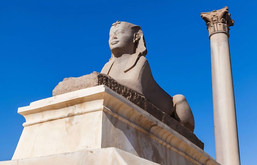 A low-angle shot of an Ancient Sphinx statue on a pedestal, with the tall, single column of Pompey Pillar rising behind it against a clear blue sky in Alexandria.