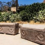 Two ancient stone sarcophagi are displayed outdoors at the Burial Catacombs Of Kom El Shoqafa, Alexandria, surrounded by desert flora, including prickly pear cacti, under a bright, sunny sky.