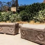 Two ancient stone sarcophagi are displayed outdoors at the Burial Catacombs Of Kom El Shoqafa, Alexandria, surrounded by desert flora, including prickly pear cacti, under a bright, sunny sky.