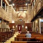 The traditional Coptic interior of the Church Of St Serge Old Cairo, showing wooden pews, brick walls, and two central columns leading to the main iconostasis.