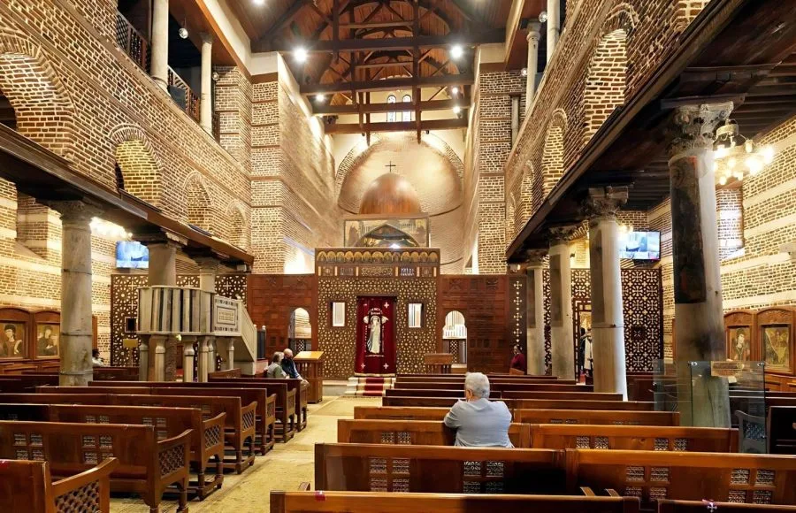 The traditional Coptic interior of the Church Of St Serge Old Cairo, showing wooden pews, brick walls, and two central columns leading to the main iconostasis.