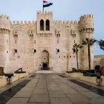 A wide, straight-on view of the massive stone Citadel Of Qaitbay Architecture in Alexandria, with its crenellated walls, central entrance tower, and the Egyptian flag flying above the battlements.