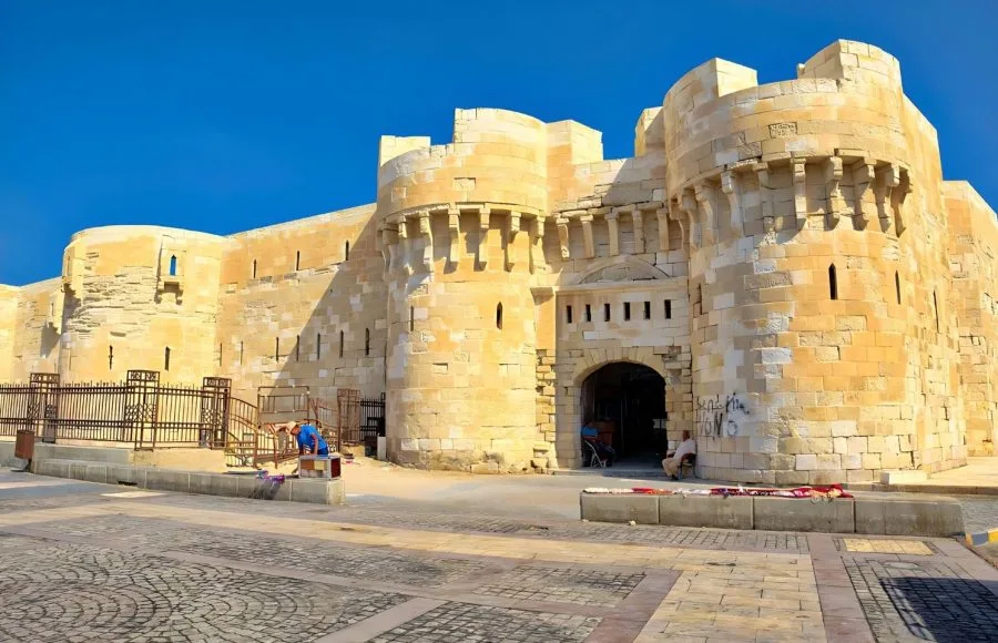 The imposing, light-colored stone fortress walls and main arched entrance of the Citadel Of Sultan Qaitbay in Alexandria, Egypt, against a bright blue sky.