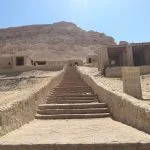 A long stone staircase leading up a sandy slope toward ancient-looking, mud-brick structures and the towering Dakrour mountain in Siwa under a bright sky.