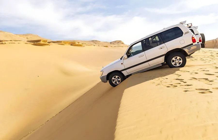 White 4x4 vehicle driving steeply down a towering sand dune at the Wadi El Hitan site.
