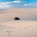 A wide, high-angle view of the gently rolling, white and golden Dunes Of The Great Sand Sea, with a black 4x4 vehicle driving across the vast, undulating landscape under a blue sky with white clouds.