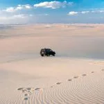 A wide, high-angle view of the gently rolling, white and golden Dunes Of The Great Sand Sea, with a black 4x4 vehicle driving across the vast, undulating landscape under a blue sky with white clouds.