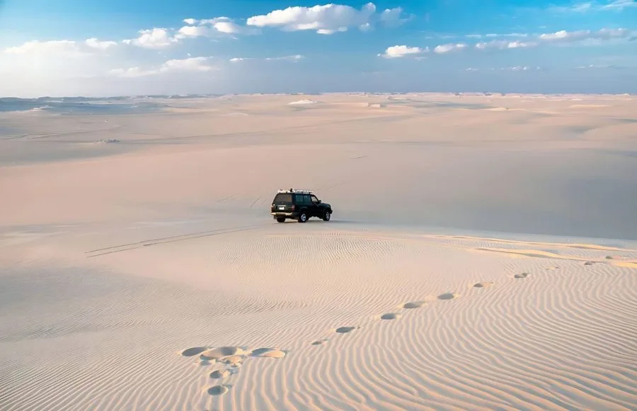 A wide, high-angle view of the gently rolling, white and golden Dunes Of The Great Sand Sea, with a black 4x4 vehicle driving across the vast, undulating landscape under a blue sky with white clouds.
