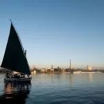 A green-sailed felucca boat on the calm Nile River with the Cairo Tower in the background.