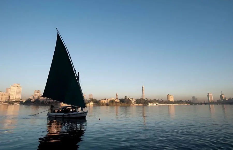A green-sailed felucca boat on the calm Nile River with the Cairo Tower in the background.