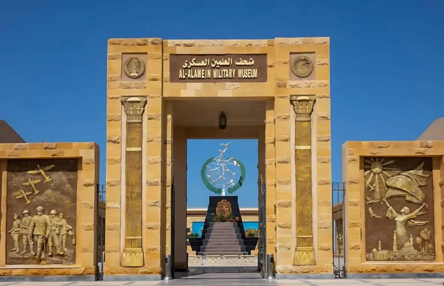 The grand, tan stone gateway to the El Alamein war memorial museum entrance, featuring Arabic and English text, pillars with historical relief carvings, and a central modern sculpture visible through the archway.