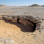 The sandy, rocky entrance to the Underground Djara Cave in the Western Desert of Egypt, surrounded by dry hills under a bright blue sky.