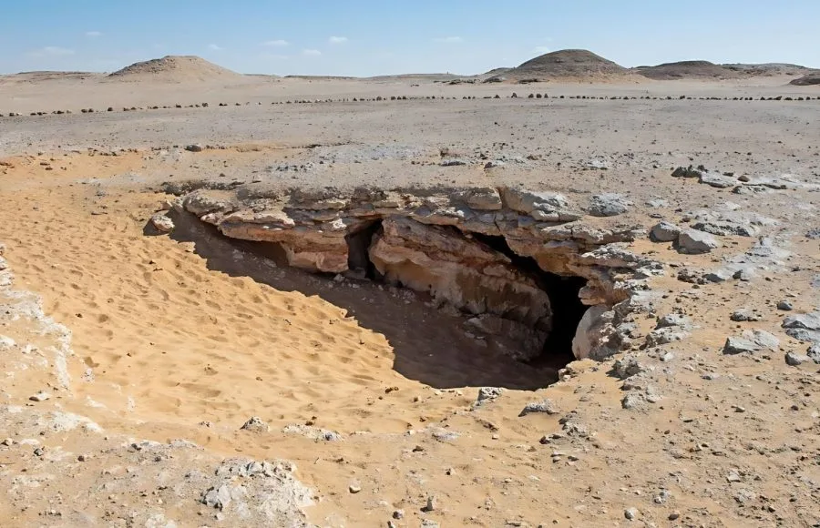 The sandy, rocky entrance to the Underground Djara Cave in the Western Desert of Egypt, surrounded by dry hills under a bright blue sky.