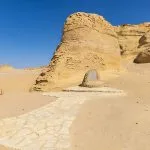 Massive, eroded yellow cliffs and a stone pathway leading to a small monument at the Wadi El Hitan site.