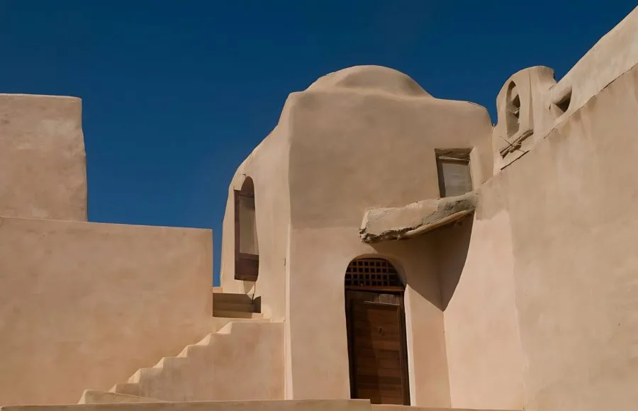 Close-up of the sunlit, geometric mud-brick architecture and staircase of the Coptic Monastery of Al-Baramous against a deep blue sky in Wadi El Natrun.