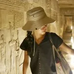 A Female Chinese tourist wearing a sun hat is explores the Temple of the goddess Hathor at Dendara, looking closely at the detailed relief carvings on the ancient Egyptian walls.