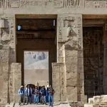 Tourists standing beneath the monumental Gateway at the Mortuary Temple of Ramesses III Medinet Habu, highlighting the massive stone entrance covered with detailed hieroglyphs and pharaonic reliefs.
