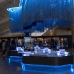 A General View Of One Of The Galleries At Grand Egyptian Museum (GEM), featuring a large, modern, blue-lit exhibit space with a unique hanging chain sculpture and groups of visitors.