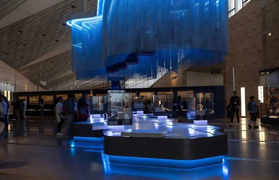 A General View Of One Of The Galleries At Grand Egyptian Museum (GEM), featuring a large, modern, blue-lit exhibit space with a unique hanging chain sculpture and groups of visitors.