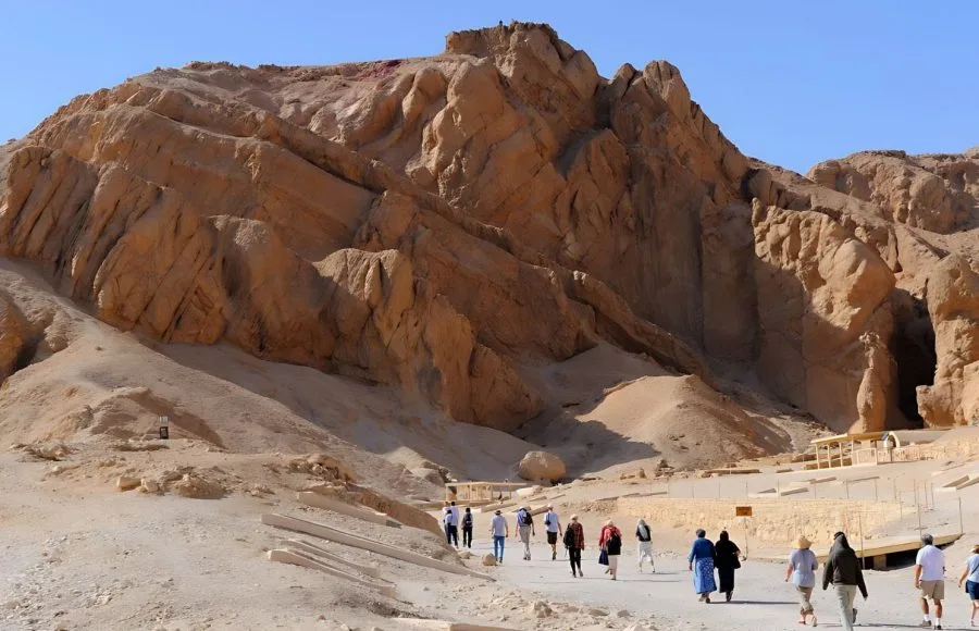 A General view of the Valley of the Queens on West Bank of River Nile at Luxor, showing the dramatic, rugged, sun-baked cliffs and a group of tourists walking on the sandy path below.