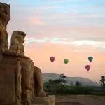 Hot air balloons floating in a pastel-colored sky over the Colossi of Memnon at sunrise, a unique sight in Luxor.