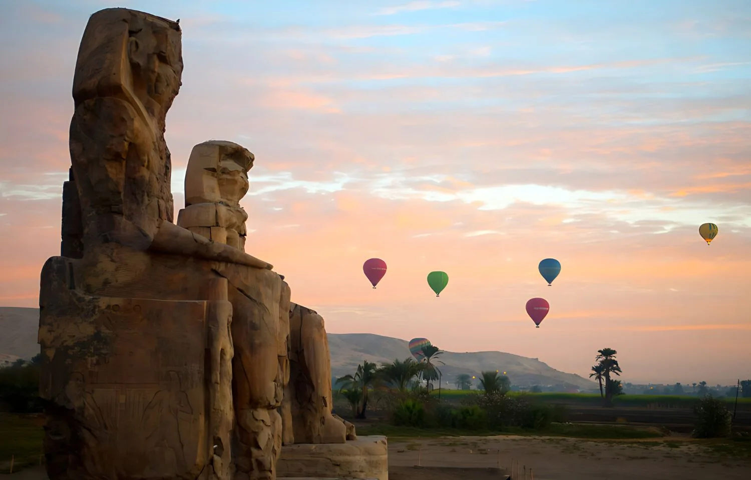 Hot air balloons floating in a pastel-colored sky over the Colossi of Memnon at sunrise, a unique sight in Luxor.