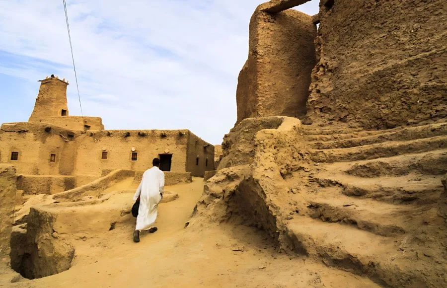 A man in a white robe walks up a winding, steep mud-brick staircase Inside Shali Fortress Siwa Oasis, surrounded by the ancient, crumbling walls and towers of the old town.
