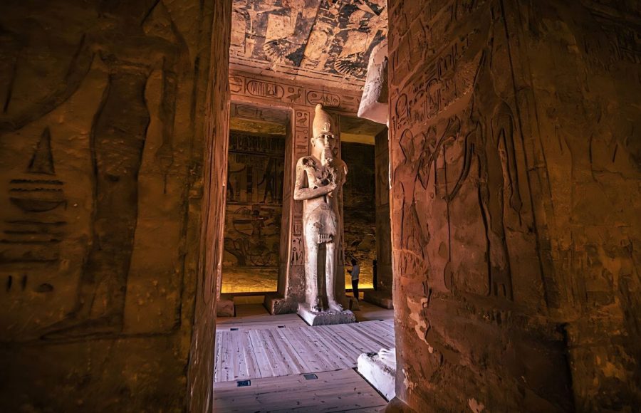 A towering Osiride pillar statue of Ramesses II deep Inside The Great Ancient Egyptian Temple Of Ramses II At Abu Simbel, illuminated by interior light and surrounded by carved wall reliefs.