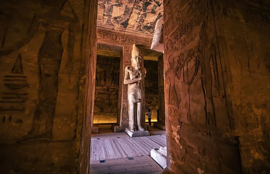 A towering Osiride pillar statue of Ramesses II deep Inside The Great Ancient Egyptian Temple Of Ramses II At Abu Simbel, illuminated by interior light and surrounded by carved wall reliefs.