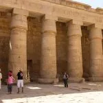 Tourists walking beneath the Large columns in the Medinet Habu temple, showing the massive, undecorated stone pillars of the peristyle court in the Mortuary Temple of Ramesses III.