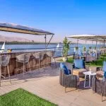 An outdoor bar and lounge area on the sundeck of the MS Historia Boutique Hotel Nile Cruise, featuring modern wicker furniture, blue cushions, and a view of the Nile River.