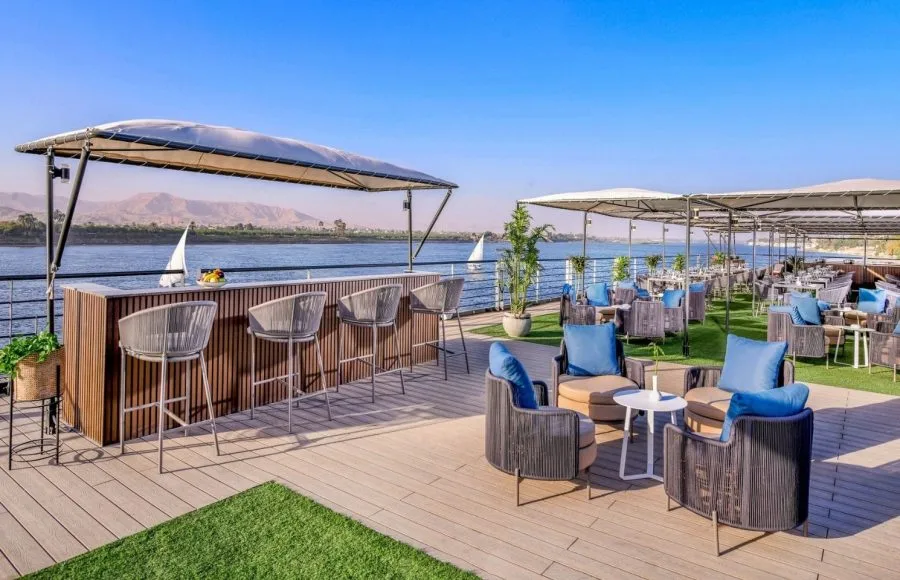 An outdoor bar and lounge area on the sundeck of the MS Historia Boutique Hotel Nile Cruise, featuring modern wicker furniture, blue cushions, and a view of the Nile River.