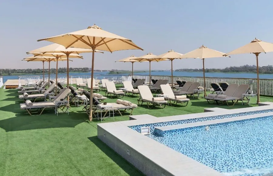 The sun deck of the Jaz Viceroy Nile Cruise, featuring a swimming pool, numerous lounge chairs, and umbrellas, with the Nile River in the background.