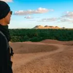 A man in a black beanie and sunglasses looking out over the dense palm groves of the Siwa Oasis from a sandy viewpoint, with rounded, low mountains visible in the distance under a blue sky.
